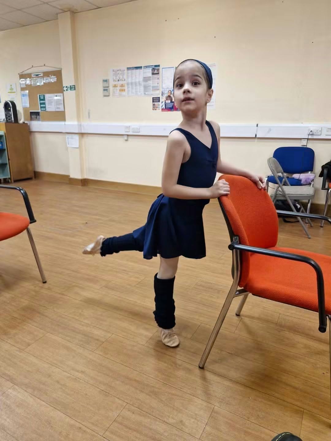 A young dancer in a navy ballet outfit practising with one leg raised beside a chair