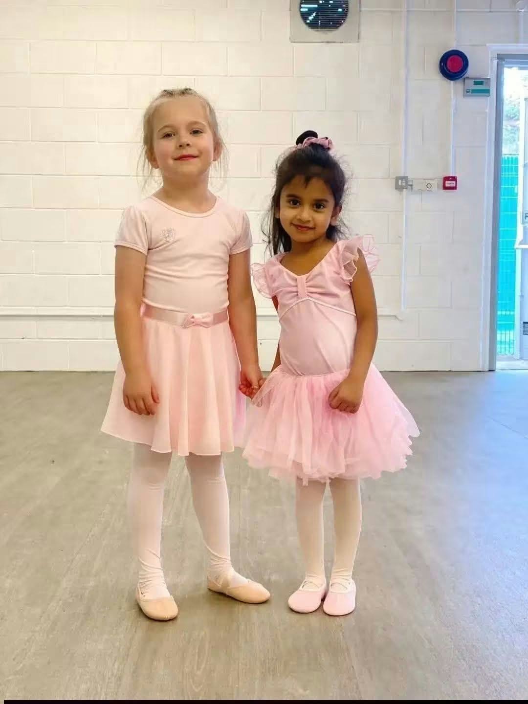 Two young dancers in pink ballet outfits standing together in a studio